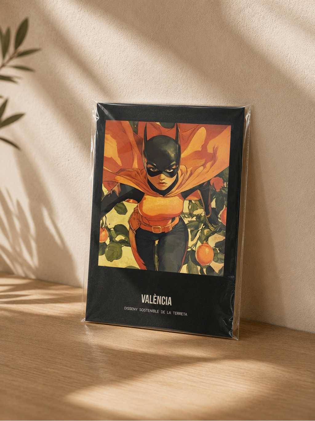 Poster València Superheróina – Mockup lámina A5 en funda transparente apoyada en madera, ilustración cómic valenciana Terreta